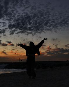 a woman standing on top of a sandy beach next to the ocean at sunset with her arms outstretched