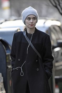 a woman with headphones is walking down the street wearing a black coat and hat