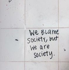 a white tiled wall with graffiti written on it and the words we blame society but we are society