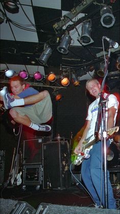 two men on stage with guitars and microphones in front of them, one man is jumping into the air