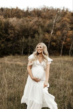 a pregnant woman in a white dress standing in tall grass with trees in the background