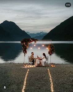 two people sitting at a table with candles in front of them on the shore of a lake