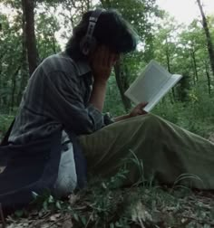 a person sitting in the woods reading a book and holding a cell phone to their ear