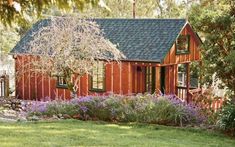 a small wooden house in the middle of a lush green yard with purple flowers and trees