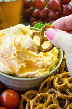 a person dipping pretzel into a bowl of dip surrounded by pretzels and grapes