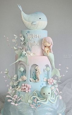 a three tiered cake with an octopus and mermaid theme on the top is decorated in pastel blue icing