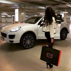 a woman standing next to a white porsche cayenne in a parking garage holding a shopping bag