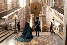 two women in blue gowns are standing at the bottom of some stairs with statues on either side