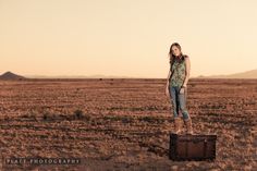 a woman standing on top of a piece of luggage in the middle of an empty field