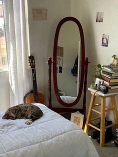 a cat laying on top of a white bed next to a tall mirror and guitar