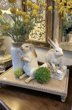 two white rabbits sitting on top of a wooden tray with flowers in the vases