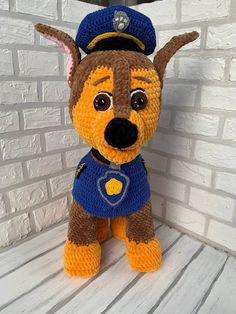 a crocheted stuffed dog wearing a police hat and blue shirt sitting in front of a brick wall