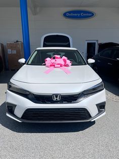 a white car with a pink bow tied to it's hood in front of a dealership