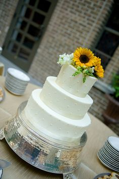 a white wedding cake with sunflowers on top sitting on a table in front of plates