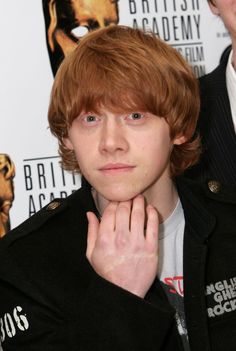 a young man with red hair wearing a black jacket and holding his hand to his face