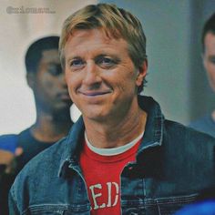 a man wearing a red shirt and jean jacket smiles at the camera while standing next to other men