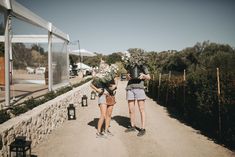 two women walking down a dirt road with flowers on their heads and one holding a plant in her hand