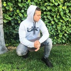 a man sitting in the grass next to a tree wearing a hoodie and looking at his cell phone