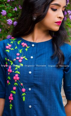 a woman wearing a blue shirt with pink flowers on the chest and shoulder, standing in front of purple flowers