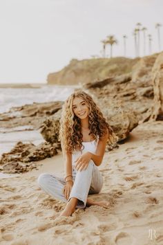 a beautiful young woman sitting on top of a sandy beach next to the ocean with palm trees