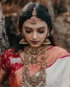 a woman wearing a red and white sari with gold jewelry on her neck, standing next to a tree