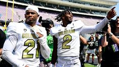 two college football players standing on the sidelines at a stadium with cameras around them