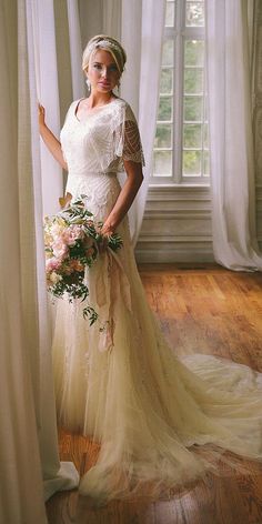 a woman standing in front of a window wearing a wedding dress and holding a bouquet