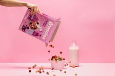 a person pouring cereal into a cup with a milk bottle next to it on a pink background