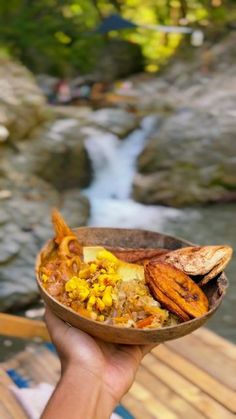 a person holding a bowl with food in it near a river and waterfall on the other side