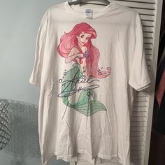 a white t - shirt with ariel from the little mermaid on it is hanging next to a hanger