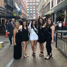 four beautiful young women standing next to each other in front of tall buildings on a city street