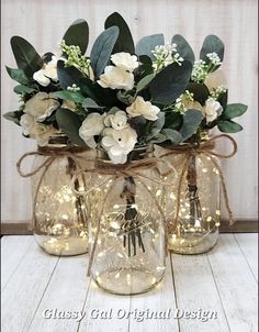 three mason jar vases with lights and flowers in them on a white wooden table