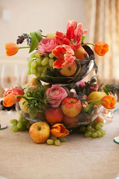 an arrangement of fruit and flowers in a bowl on a table with wine glassware