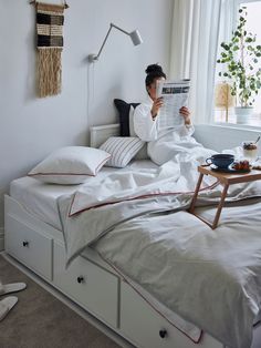 a person sitting on a bed reading a newspaper in a bedroom with white walls and flooring