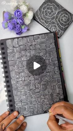 two hands are holding pens and writing on a notebook with an intricate pattern in the background