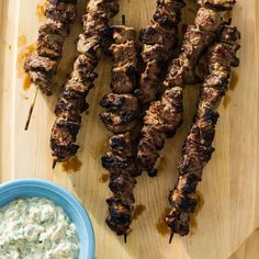 several skewers of meat on a wooden cutting board next to a bowl of dip