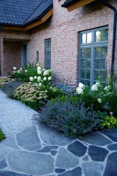 a stone walkway leads to a brick house with white flowers in the foreground and green grass on the other side