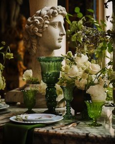 a table topped with green glass vases filled with flowers and greenery next to a statue