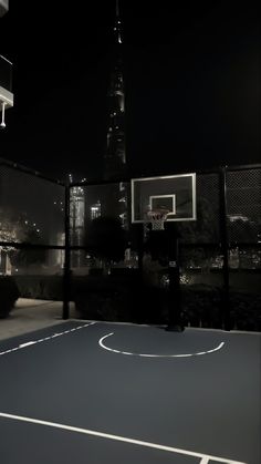 an empty basketball court in front of a cityscape at night with the lights on