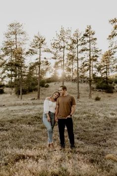 a man and woman standing in the middle of a field with pine trees behind them