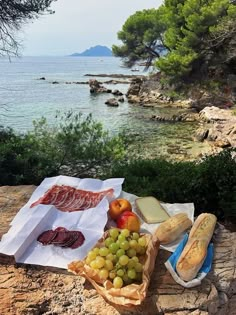 an assortment of cheeses, fruit and bread on a rock near the water's edge