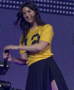 a woman in a yellow shirt and black skirt on stage with her arms out to the side