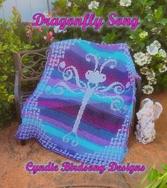 a crocheted dragonfly blanket sitting on top of a bench next to flowers