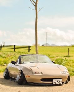 a tan sports car parked in front of a tree on a dirt road near a field