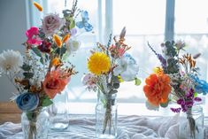 three vases filled with different colored flowers on a white table cloth next to a window