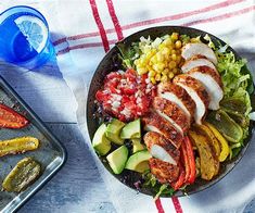 a salad with chicken, avocado, corn and tomatoes on a plate next to a tray of pickles