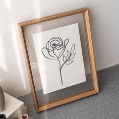 a black and white drawing of a rose in a wooden frame on the floor next to a wall