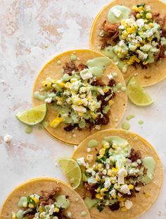 three tacos filled with meat and vegetables on top of a white countertop next to lime wedges