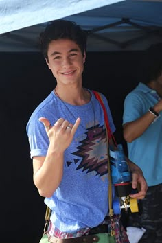 a young man standing under a tent holding a skateboard and giving the peace sign