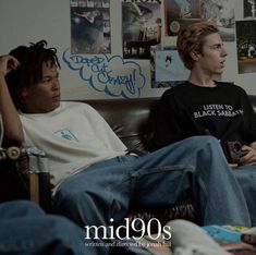 two young men sitting on a couch in front of a wall covered with pictures and posters